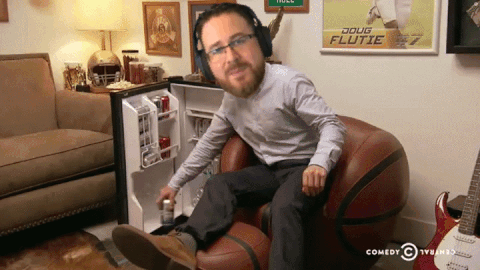 A Man Wearing Headphones Sits In A Chair In Front Of A Doug Flutie Poster GIF
