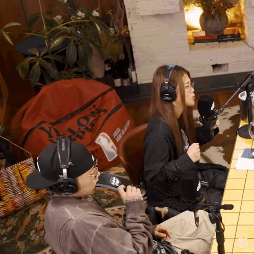 A Man Wearing Headphones Sits Next To A Woman Wearing Headphones In Front Of A Red Bag That Says Lion GIF