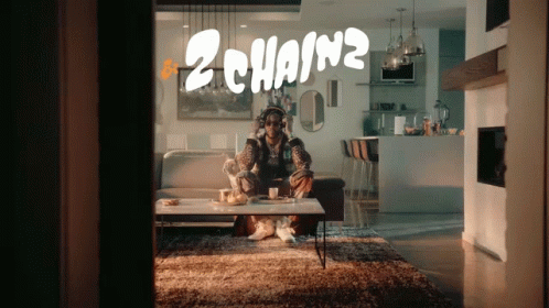 A Man Wearing Headphones Sits On A Couch In A Living Room With 2 Chainz Written On The Wall Behind Him GIF