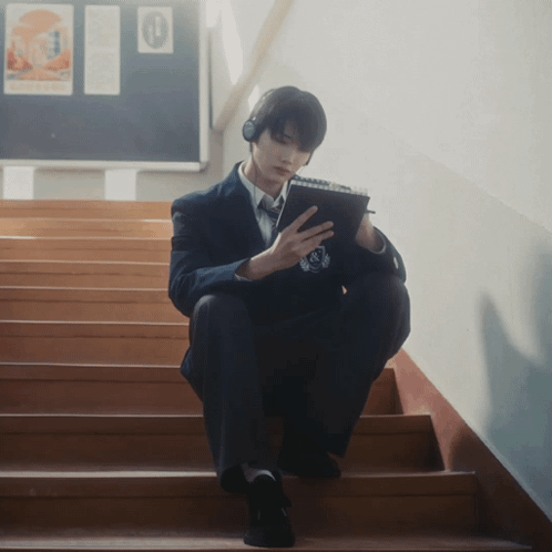A Man Wearing Headphones Sits On A Set Of Stairs Looking At His Phone GIF