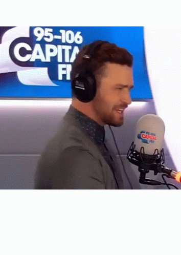 A Man Wearing Headphones Stands In Front Of A Microphone That Says 95-106 Capital Fm On It GIF