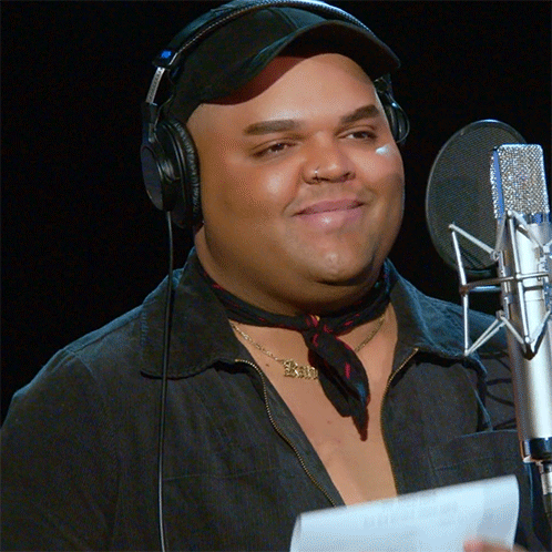A Man Wearing Headphones Stands In Front Of A Microphone With A Necklace That Says Exi GIF