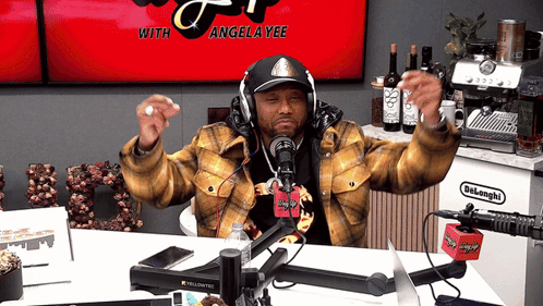 A Man Wearing Headphones Stands In Front Of A Microphone With Angela Yee On The Screen GIF