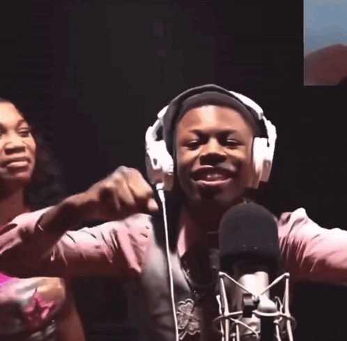 A Man Wearing Headphones Stands In Front Of A Microphone With His Hands In The Air GIF