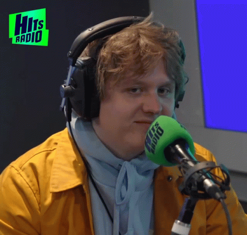 A Man Wearing Headphones Stands In Front Of A Microphone With The Words Hits Radio In The Background GIF