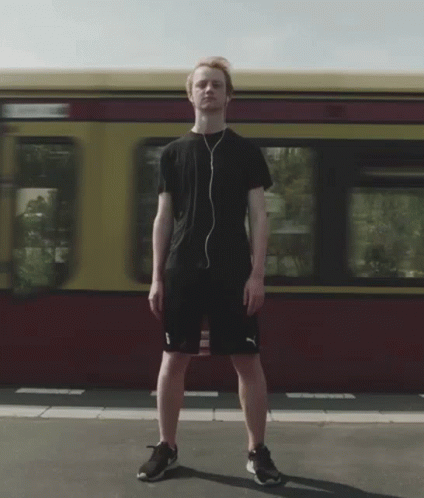 A Man Wearing Headphones Stands In Front Of A Red And Yellow Train GIF