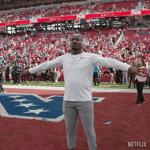 A Man Wearing Headphones Stands On A Football Field With His Arms Outstretched In Front Of A Us Bank Banner GIF