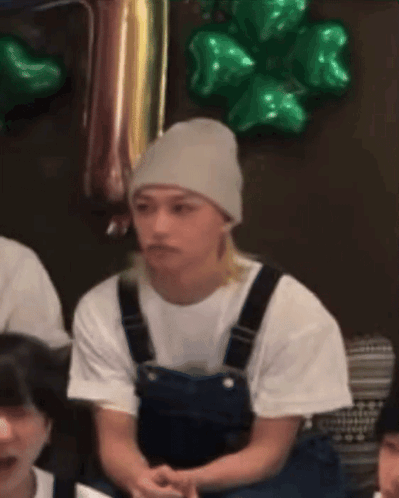 A Man Wearing Overalls And A Beanie Is Sitting In Front Of Green Balloons .. GIF