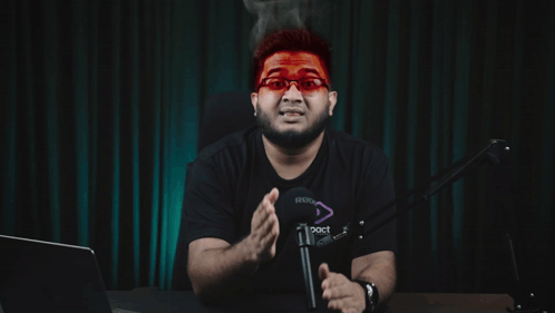A Man Wearing Red Glasses Is Sitting In Front Of A Microphone With A Foreign Language Behind Him GIF