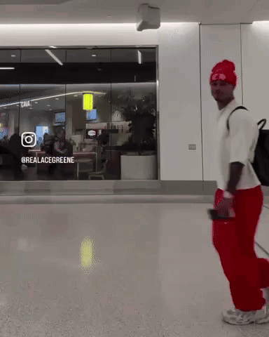 A Man Wearing Red Pants And A White Shirt Is Walking In Front Of A Glass Window That Says @Realacegreene GIF