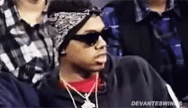 A Man Wearing Sunglasses , A Bandana And A Necklace Is Sitting In A Crowd Of People . GIF