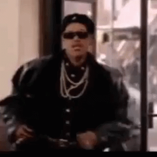 A Man Wearing Sunglasses , A Hat , And A Necklace Is Standing In Front Of A Glass Door . GIF