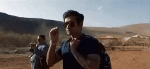 A Man Wearing Sunglasses And A Backpack Is Walking Through A Desert . GIF