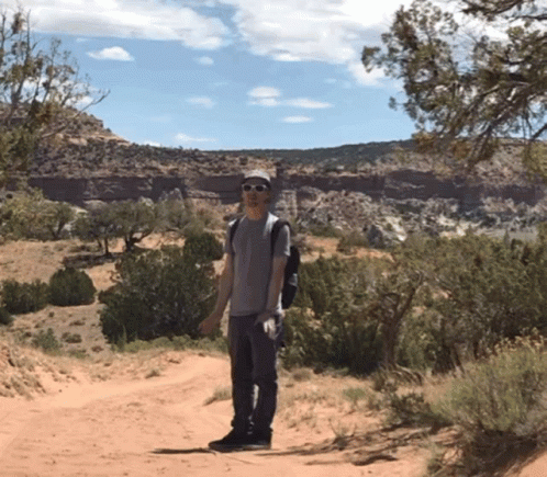 A Man Wearing Sunglasses And A Backpack Stands On A Dirt Road In The Desert GIF