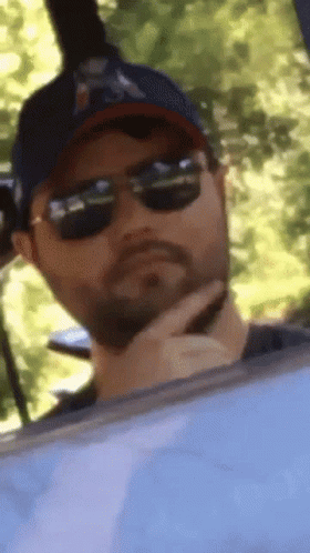 A Man Wearing Sunglasses And A Baseball Cap Is Looking Out Of A Window GIF