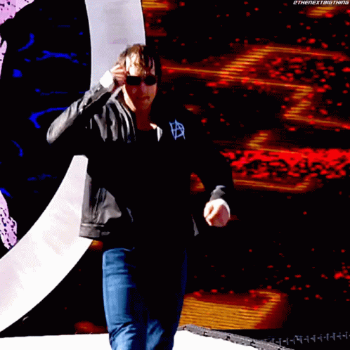 A Man Wearing Sunglasses And A Black Jacket Stands In Front Of A Large Screen That Says Thenextbigthing GIF
