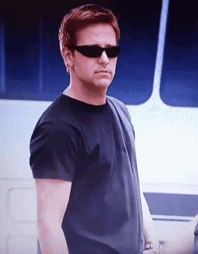 A Man Wearing Sunglasses And A Black Shirt Is Standing In Front Of A Boat GIF