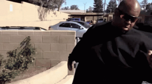 A Man Wearing Sunglasses And A Black Shirt Stands In Front Of A Parking Lot GIF