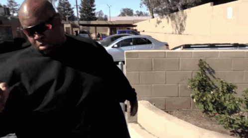 A Man Wearing Sunglasses And A Black Shirt Stands In Front Of A Parking Lot GIF