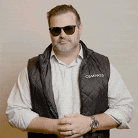 A Man Wearing Sunglasses And A Black Vest With The Word Compass On It GIF