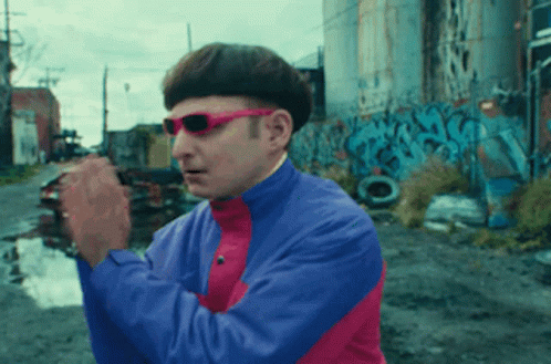 A Man Wearing Sunglasses And A Blue And Pink Jacket Is Standing In Front Of Graffiti GIF