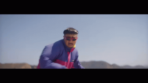 A Man Wearing Sunglasses And A Blue Jacket Is Standing In The Desert GIF