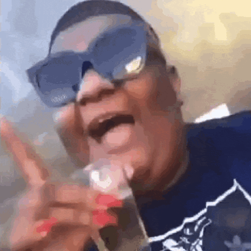 A Man Wearing Sunglasses And A Blue Shirt Is Holding A Glass In His Hand . GIF