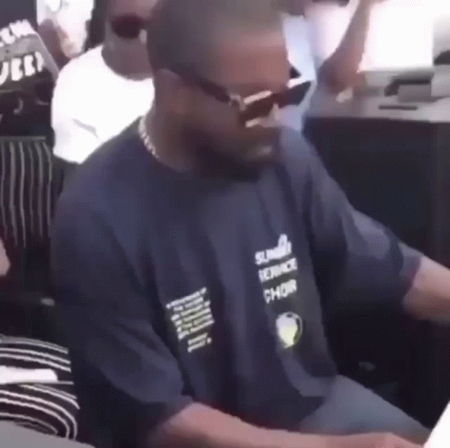A Man Wearing Sunglasses And A Blue Shirt Is Sitting At A Table In A Crowd . GIF