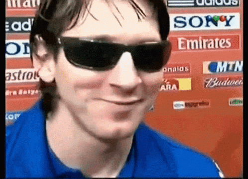 A Man Wearing Sunglasses And A Blue Shirt Is Smiling In Front Of A Sign That Says Sony Emirates GIF