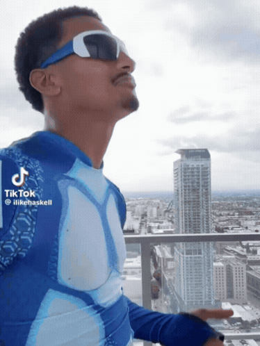 A Man Wearing Sunglasses And A Blue Shirt Is Standing On A Balcony With A City In The Background GIF