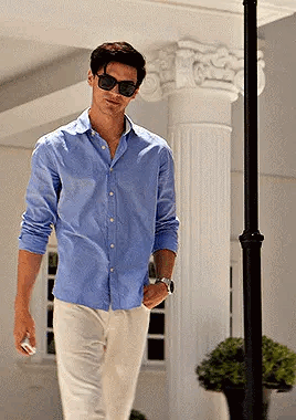 A Man Wearing Sunglasses And A Blue Shirt Is Walking Down A Street GIF