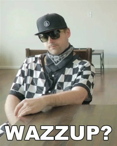 A Man Wearing Sunglasses And A Checkered Shirt Is Sitting At A Table With The Words Wazzup On The Table GIF