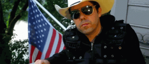 A Man Wearing Sunglasses And A Cowboy Hat Stands In Front Of An American Flag GIF