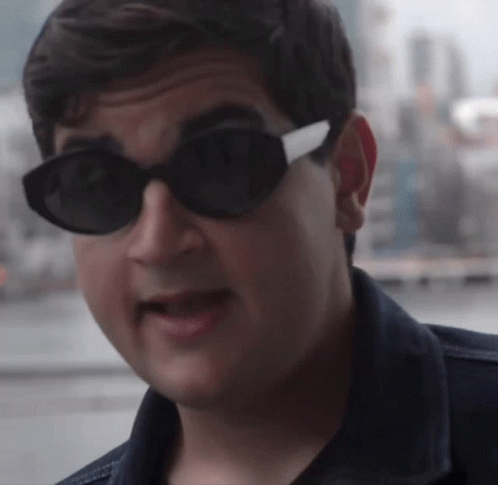 A Man Wearing Sunglasses And A Denim Jacket Looks At The Camera GIF