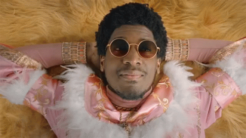 A Man Wearing Sunglasses And A Feather Boa Is Laying Down GIF