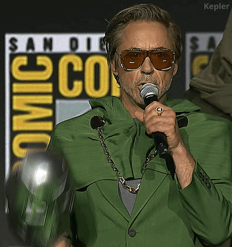 A Man Wearing Sunglasses And A Green Jacket Is Holding A Microphone In Front Of A San Diego Comic Con Sign GIF