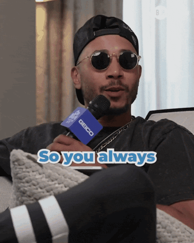 A Man Wearing Sunglasses And A Hat Is Holding A Microphone That Says Geico On It GIF