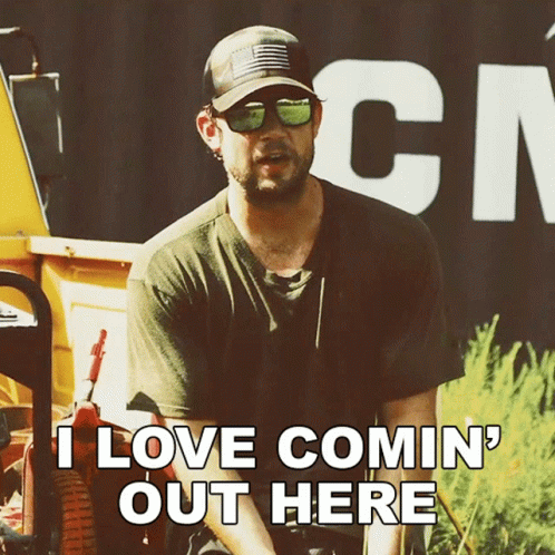 A Man Wearing Sunglasses And A Hat Is Sitting In Front Of A Sign That Says " I Love Comin ' Out Here " GIF