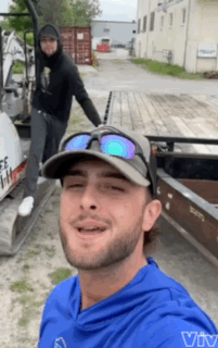 A Man Wearing Sunglasses And A Hat Is Smiling In Front Of A Boat GIF