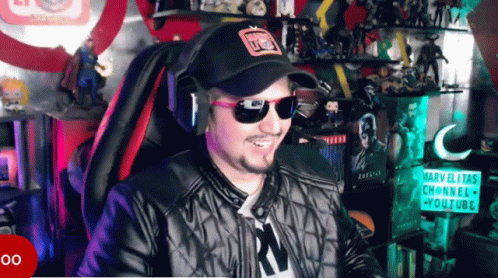 A Man Wearing Sunglasses And A Hat Is Smiling In Front Of A Sign That Says Marvelitas Channel GIF