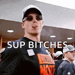 A Man Wearing Sunglasses And A Hat Is Smoking A Cigarette And Says " Sup Bitches " . GIF