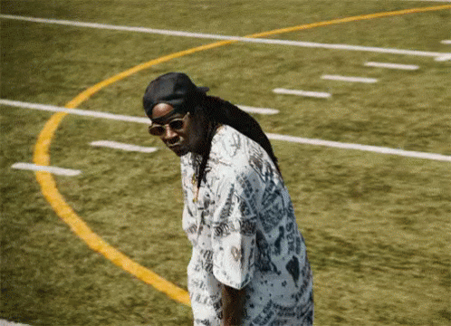 A Man Wearing Sunglasses And A Hat Is Standing On A Field GIF