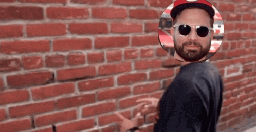 A Man Wearing Sunglasses And A Hat Stands In Front Of A Red Brick Wall GIF