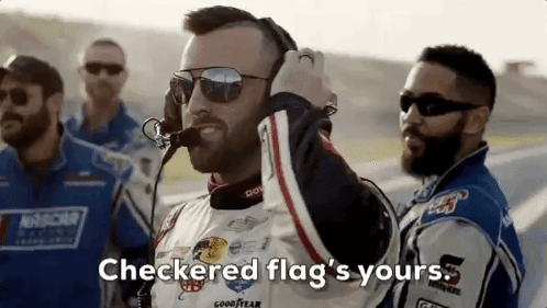 A Man Wearing Sunglasses And A Headset Is Standing Next To A Group Of Race Car Drivers On A Race Track . GIF