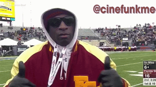 A Man Wearing Sunglasses And A Hooded Jacket Giving A Thumbs Up In Front Of A Football Field GIF