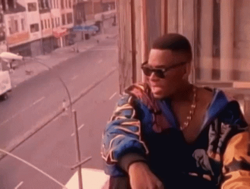 A Man Wearing Sunglasses And A Jacket Is Sitting On A Balcony Overlooking A Street . GIF