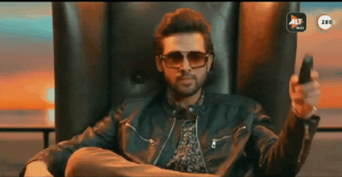 A Man Wearing Sunglasses And A Leather Jacket Is Sitting In A Chair Holding A Remote Control GIF