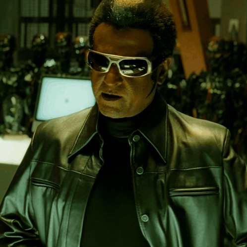 A Man Wearing Sunglasses And A Leather Jacket Stands In Front Of A Computer Monitor GIF