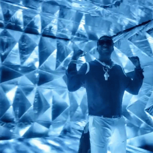 A Man Wearing Sunglasses And A Necklace Is Dancing In Front Of A Blue Wall GIF