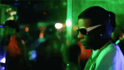 A Man Wearing Sunglasses And A Necklace Is Standing In A Crowded Room . GIF